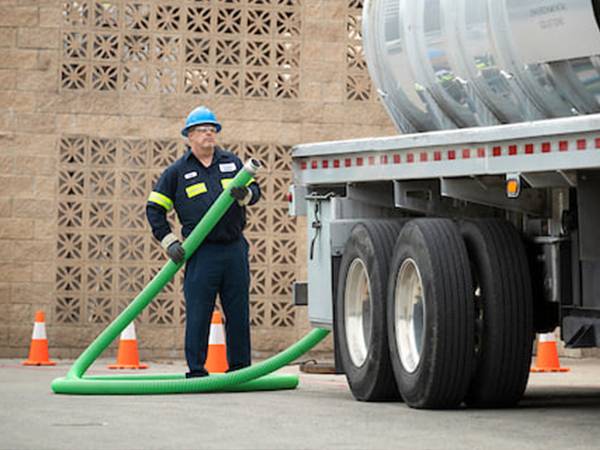 A worker standing next to a sliver tanker holds a green chemical hose.